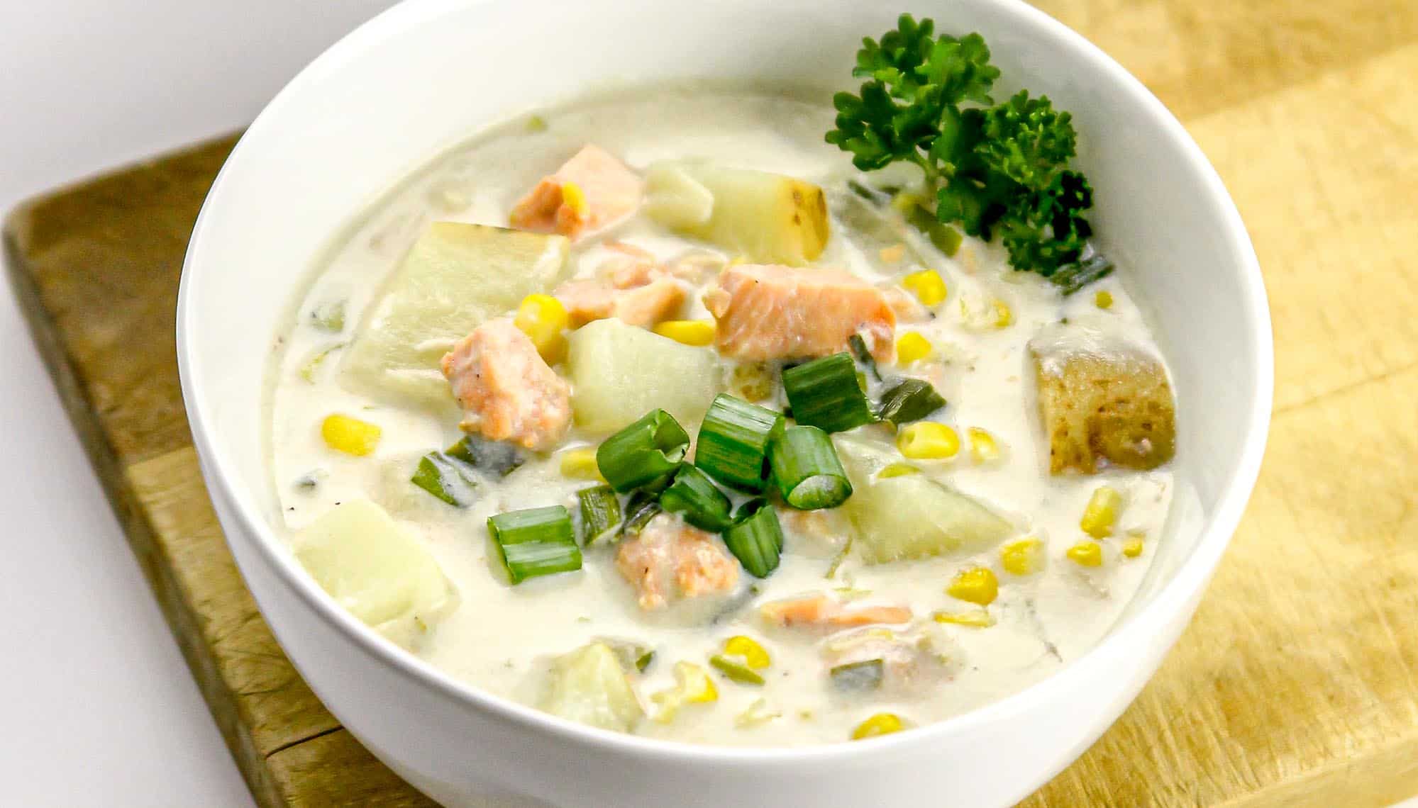 Creamy fish chowder with chunks of potato, corn, green onions, and garnished with parsley in a white bowl on a wooden board.