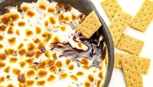 Toasted marshmallows and melted chocolate in a skillet with graham crackers beside it on a white surface.