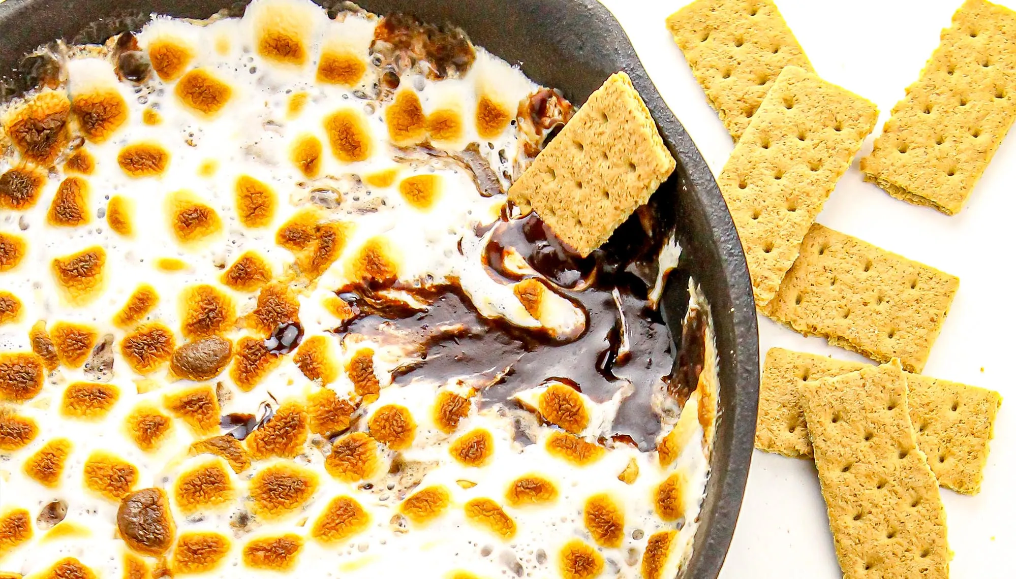 Toasted marshmallows and melted chocolate in a skillet with graham crackers beside it on a white surface.