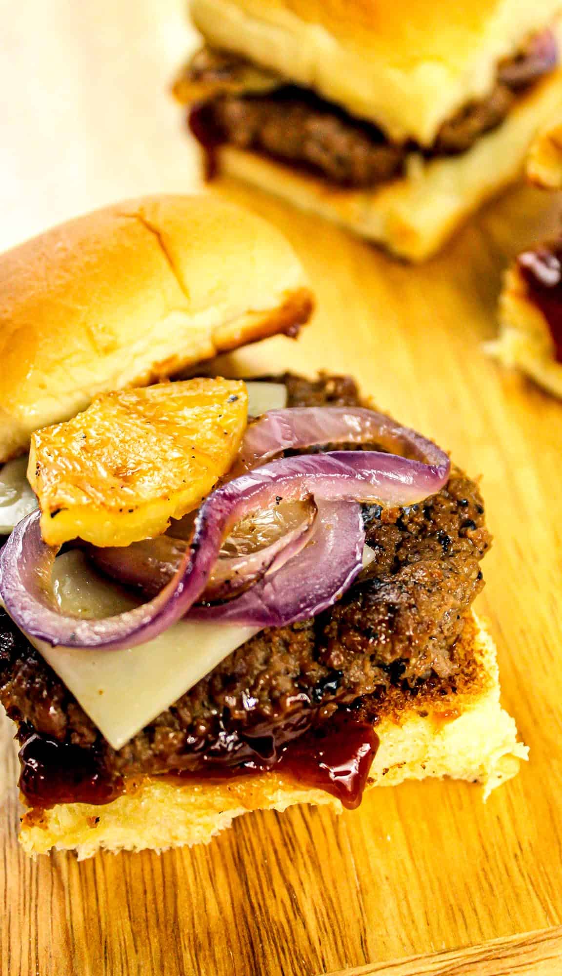 Hamburger with grilled pineapple, red onion, Swiss cheese, and barbecue sauce on a toasted bun on a wooden surface.