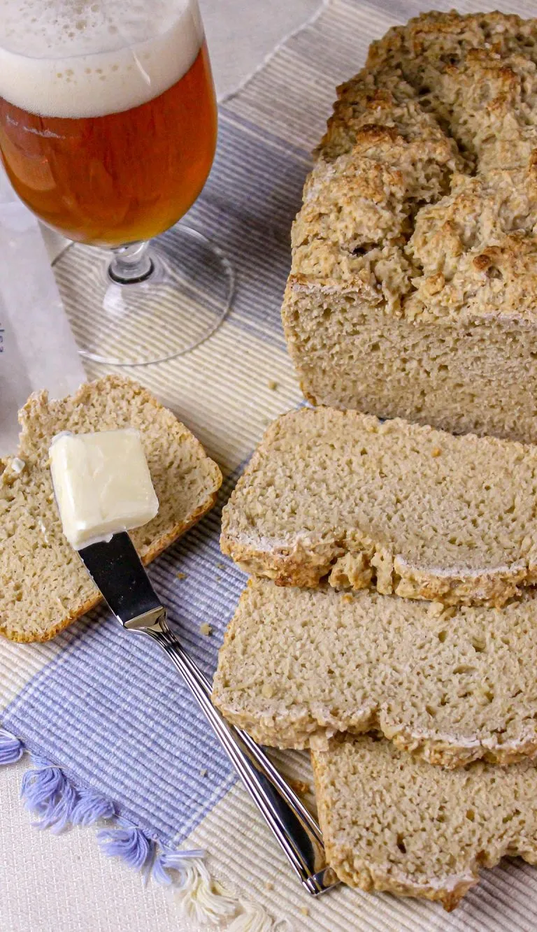 Homemade soda bread with sliced pieces, a buttered slice with a butter knife, and a glass of amber beer on a tablecloth.