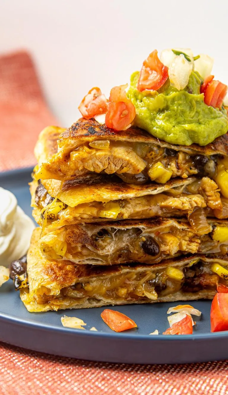 Stack of chicken, corn, black bean quesadilla slices topped with guacamole, diced tomatoes, and onions on blue plate with sour cream on the side.