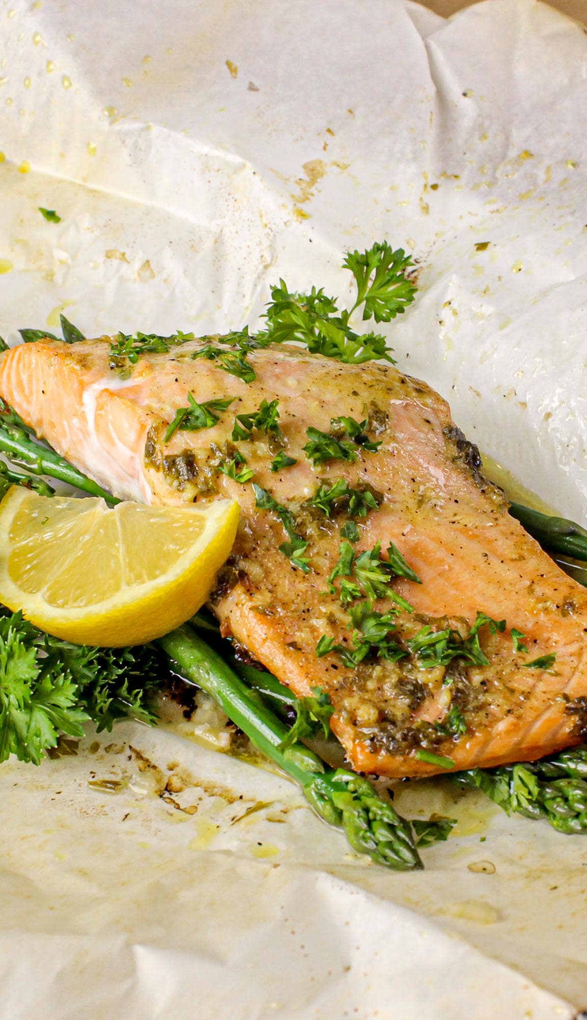Baked salmon fillet garnished with herbs on asparagus spears with a lemon wedge and parsley on parchment paper.