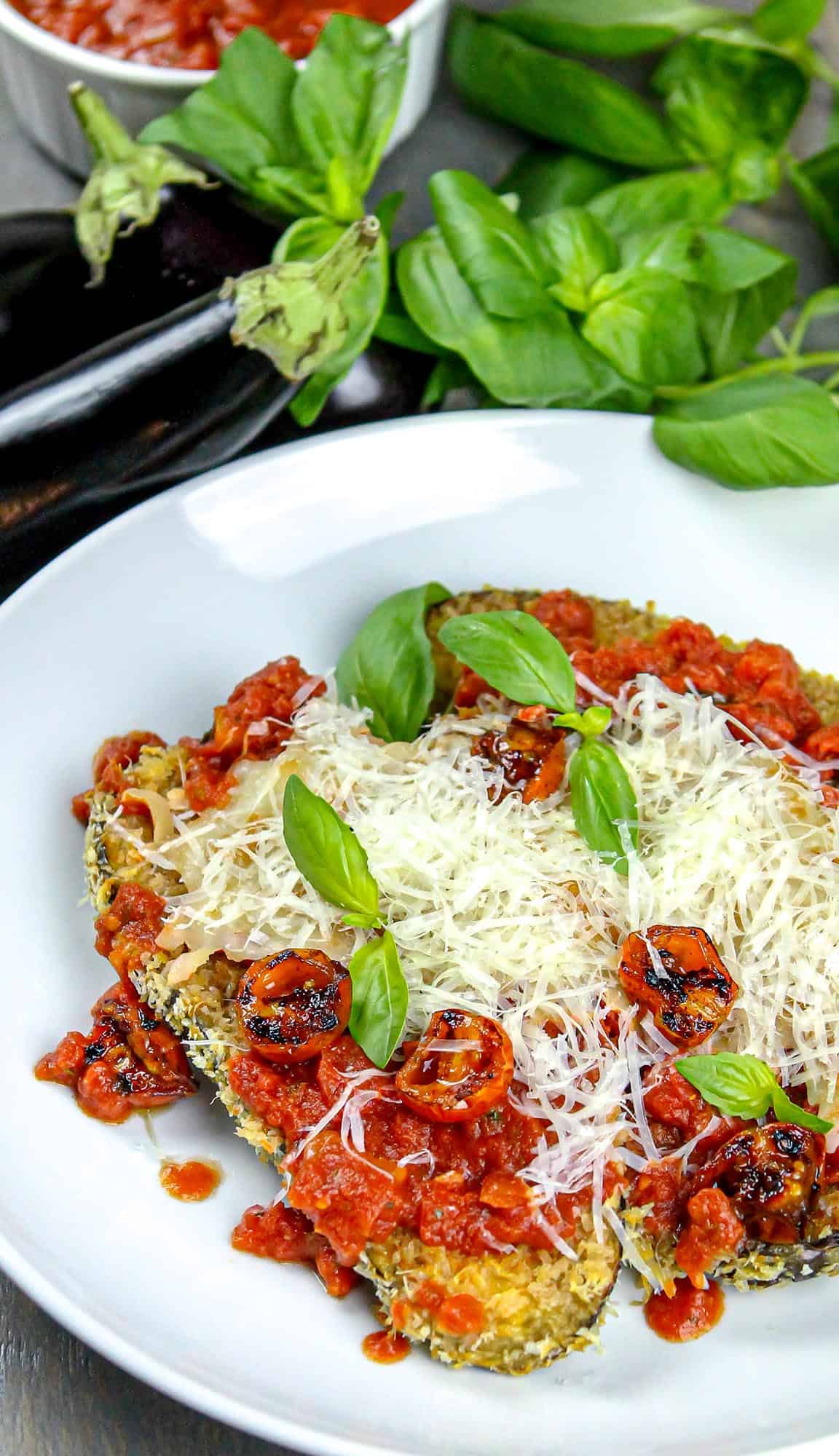 Breaded eggplant slices topped with tomato sauce, grilled cherry tomatoes, shredded cheese, and fresh basil leaves on a white plate.