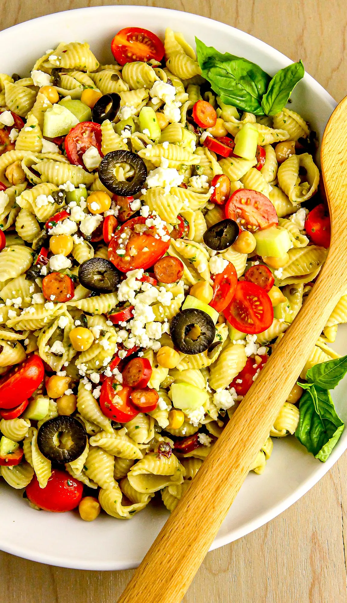 Pasta salad with shell pasta, cherry tomatoes, black olives, chickpeas, cucumber, feta cheese, basil leaves, and wooden spoon on a white plate.