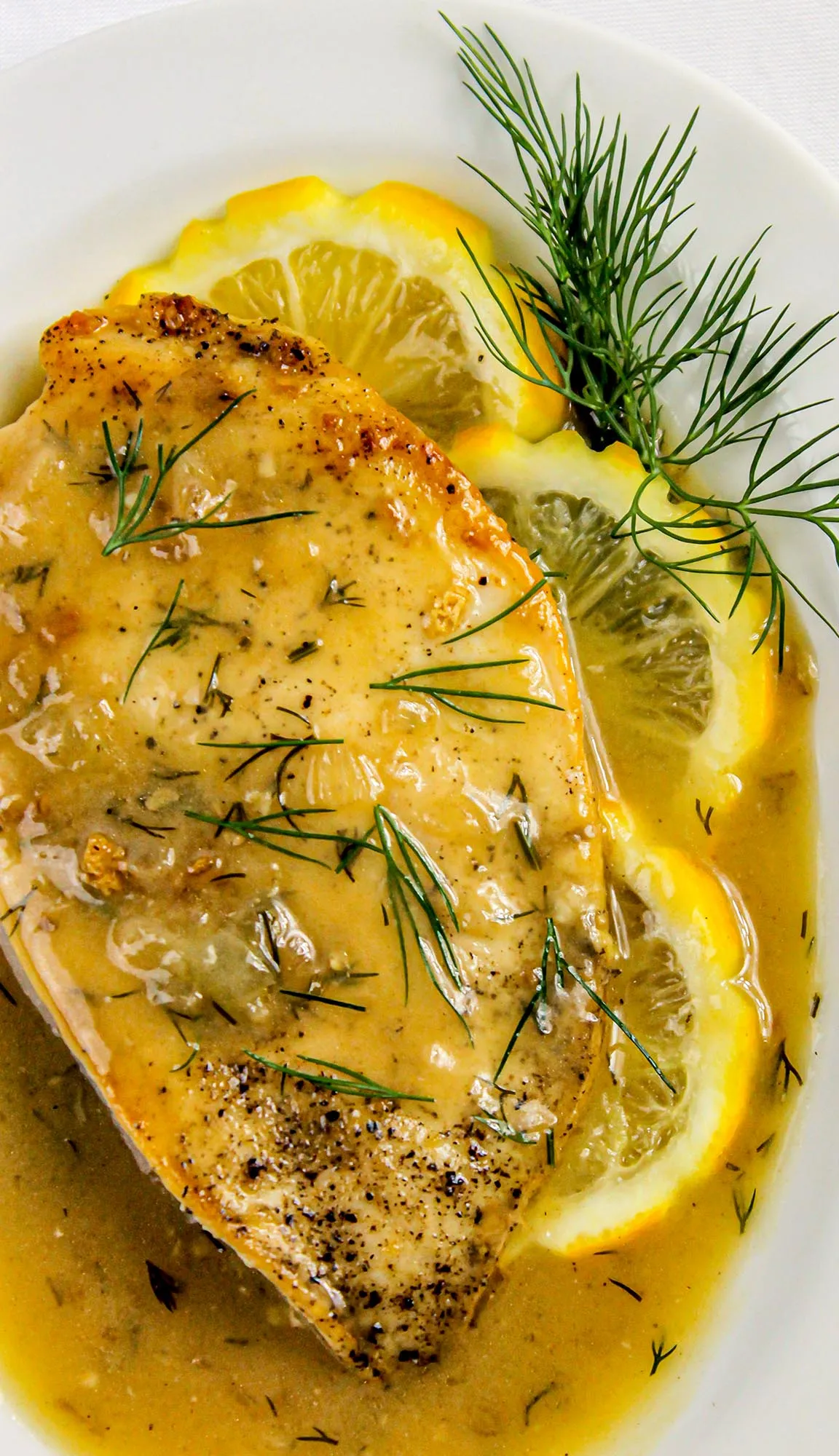Chicken breast in lemon dill sauce garnished with fresh dill and lemon slices on a white plate.