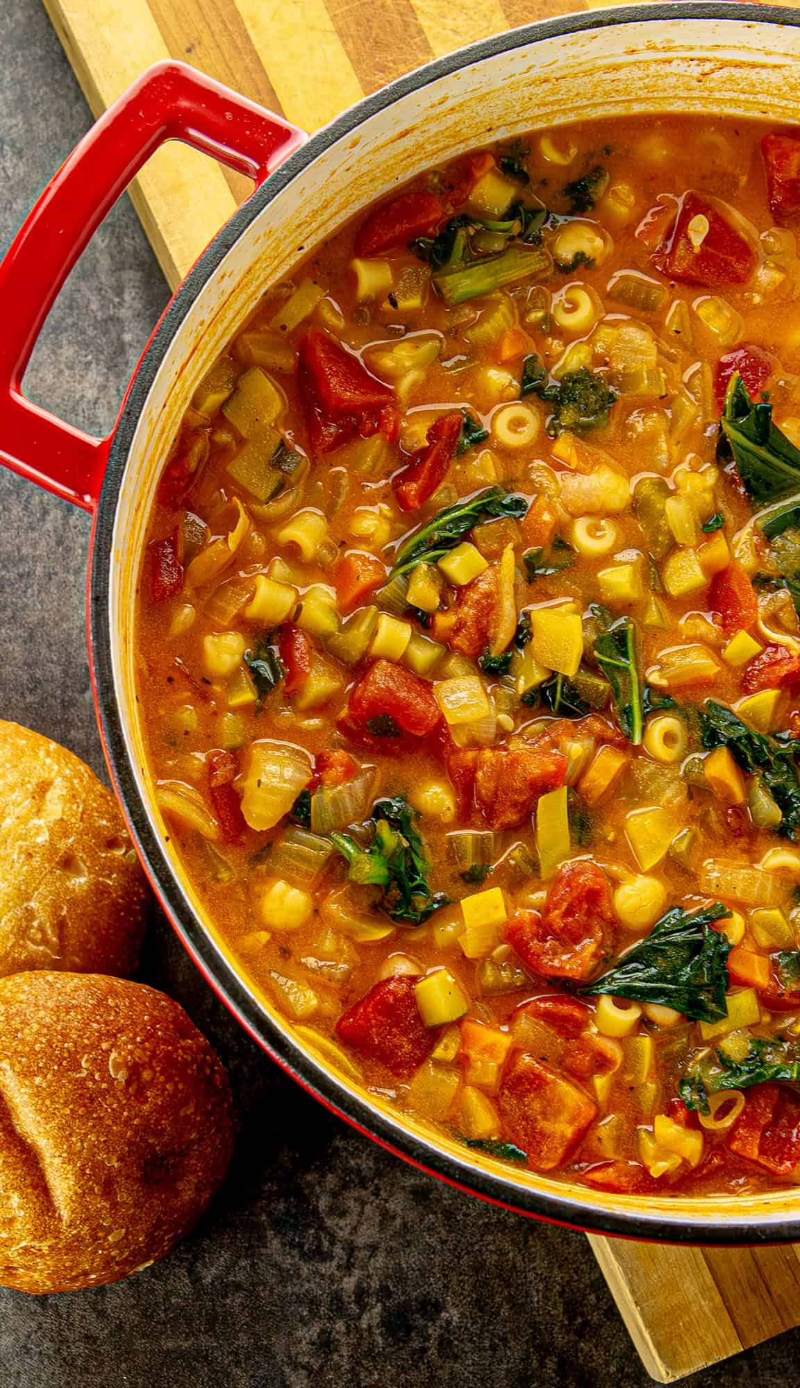 Red pot with vegetable minestrone soup containing tomatoes, spinach, pasta, and other diced vegetables, with two bread rolls on the side.