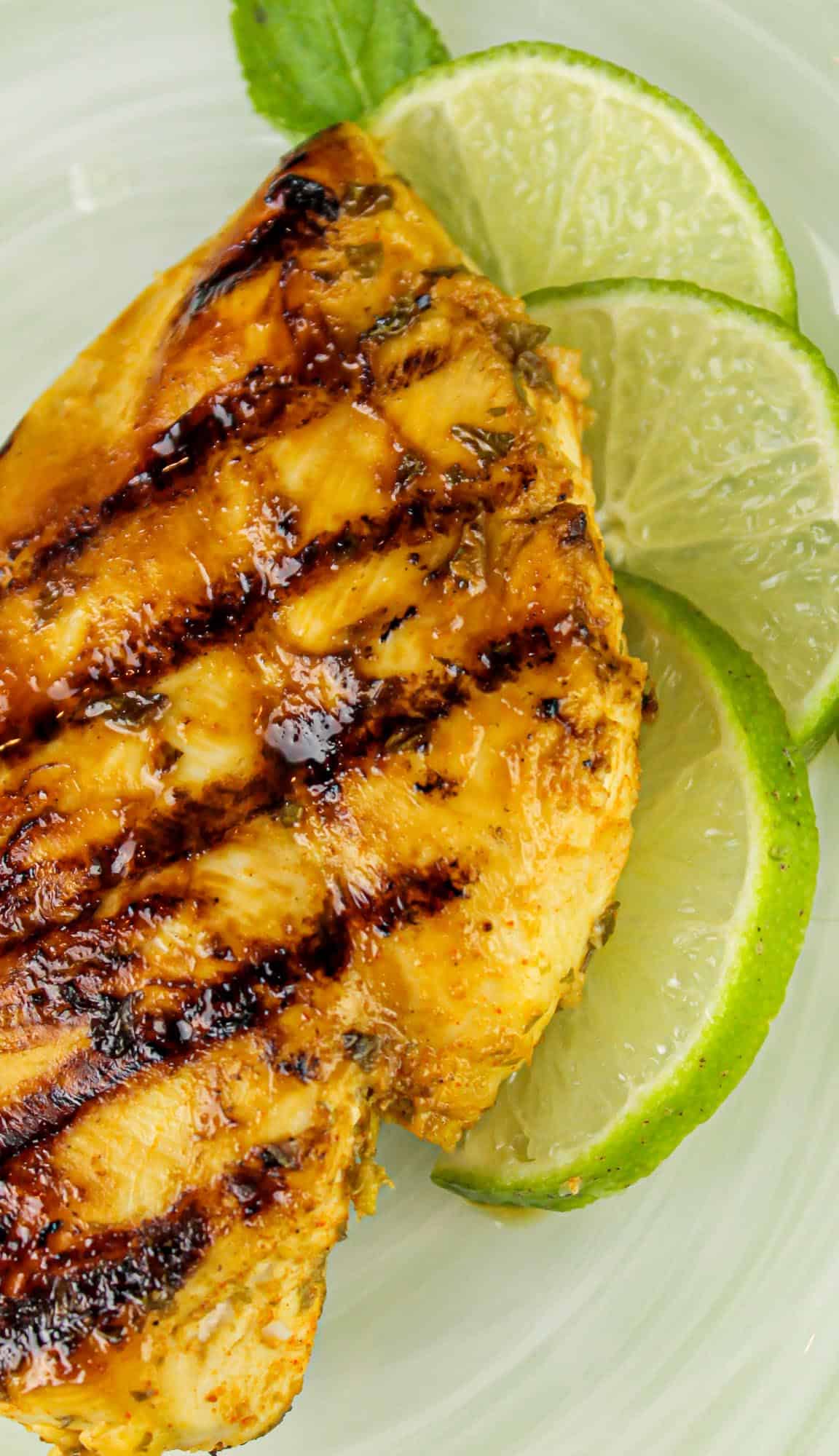 Grilled chicken breast with char marks, garnished with lime slices and a mint leaf on a white plate.