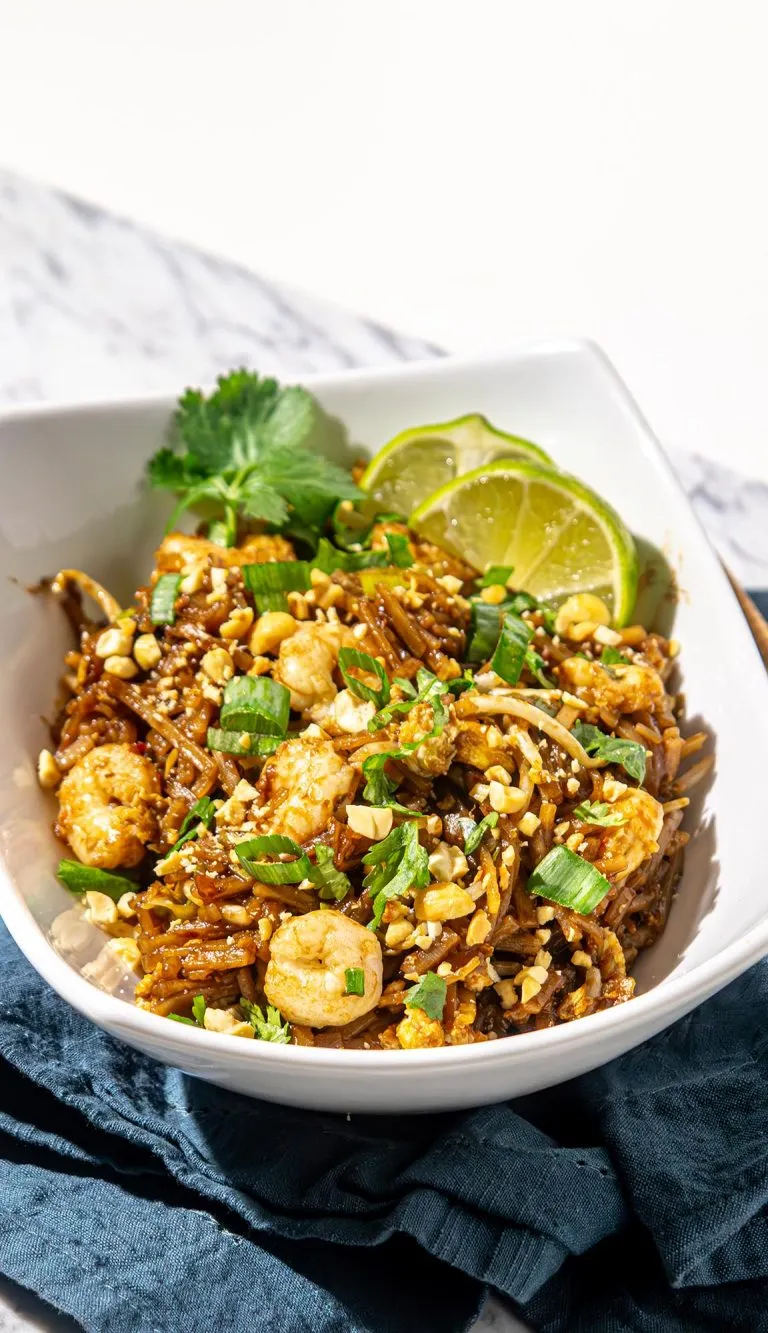 Shrimp Pad Thai noodles garnished with crushed peanuts, green onions, cilantro, and lime wedges in a white bowl on a dark cloth.