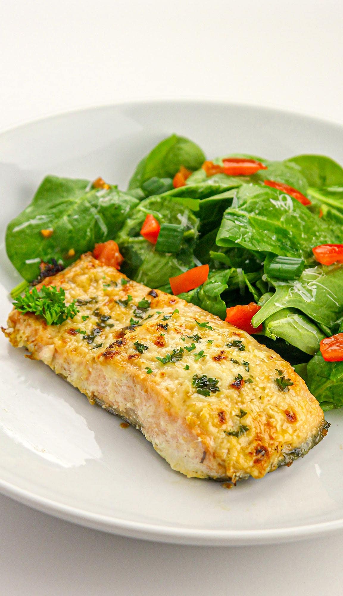 Baked salmon fillet garnished with herbs served with fresh spinach salad topped with diced tomatoes and green onions on a white plate.