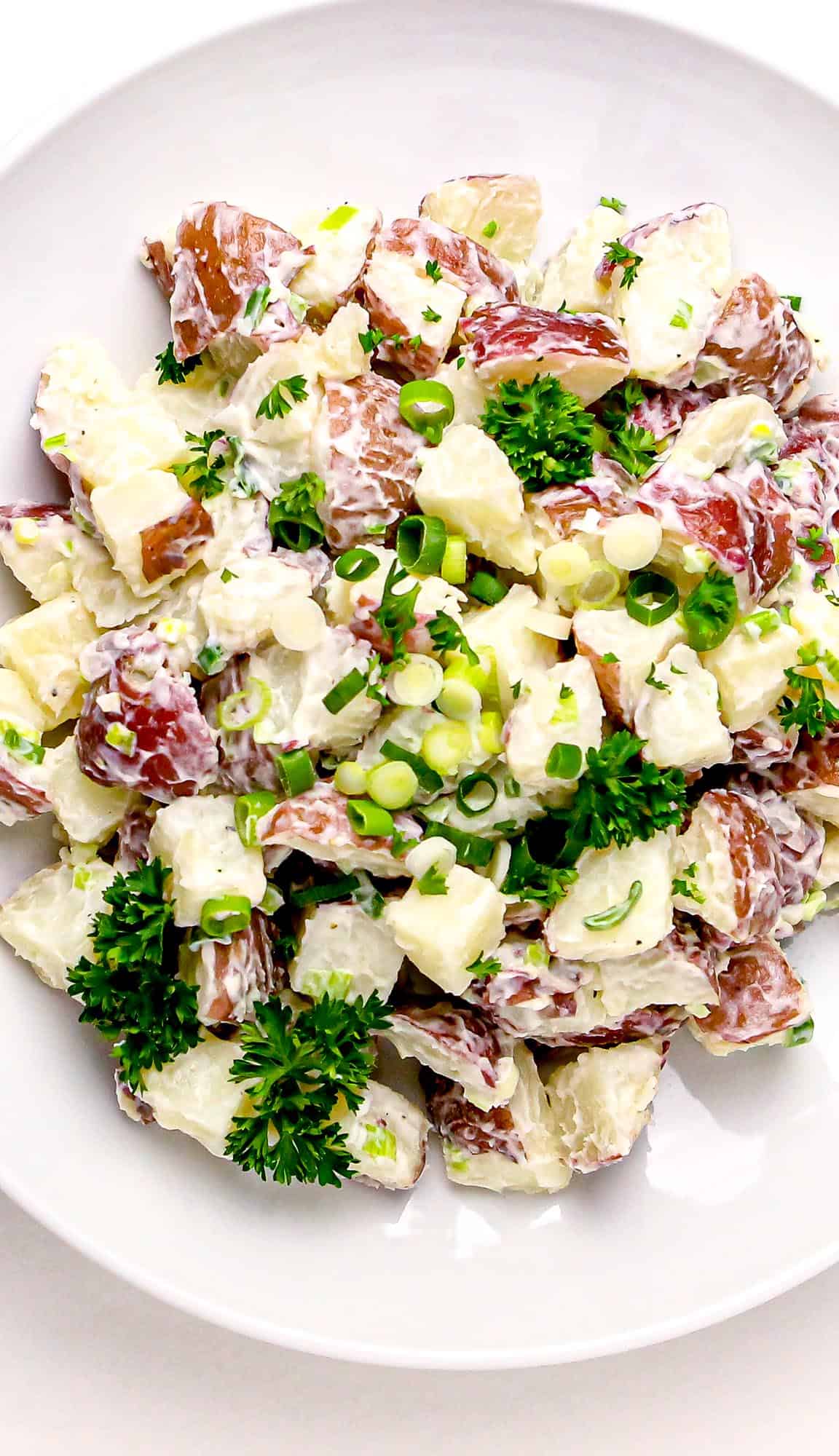 Red-skinned potato salad mixed with creamy dressing, chopped green onions, and fresh parsley in a white bowl.