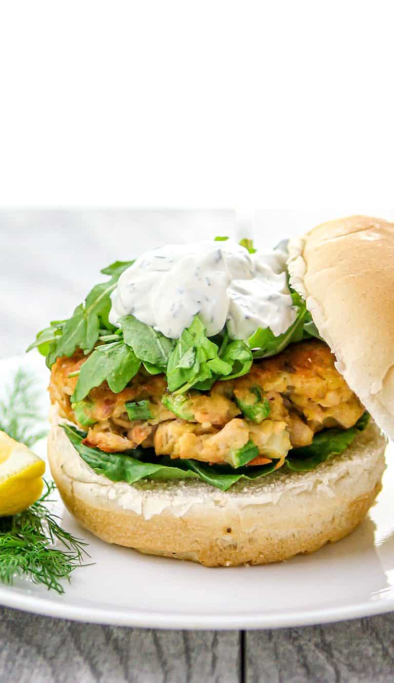 Chickpea patty sandwich with greens and creamy herb sauce on a bun, served on a white plate with lemon wedge and dill garnish.
