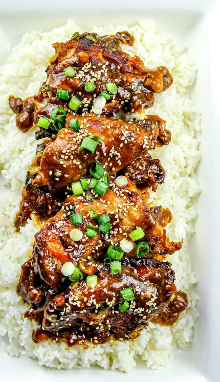 Chicken thighs glazed with dark sauce, sprinkled with green onions and sesame seeds, served on a bed of white rice in a white dish.