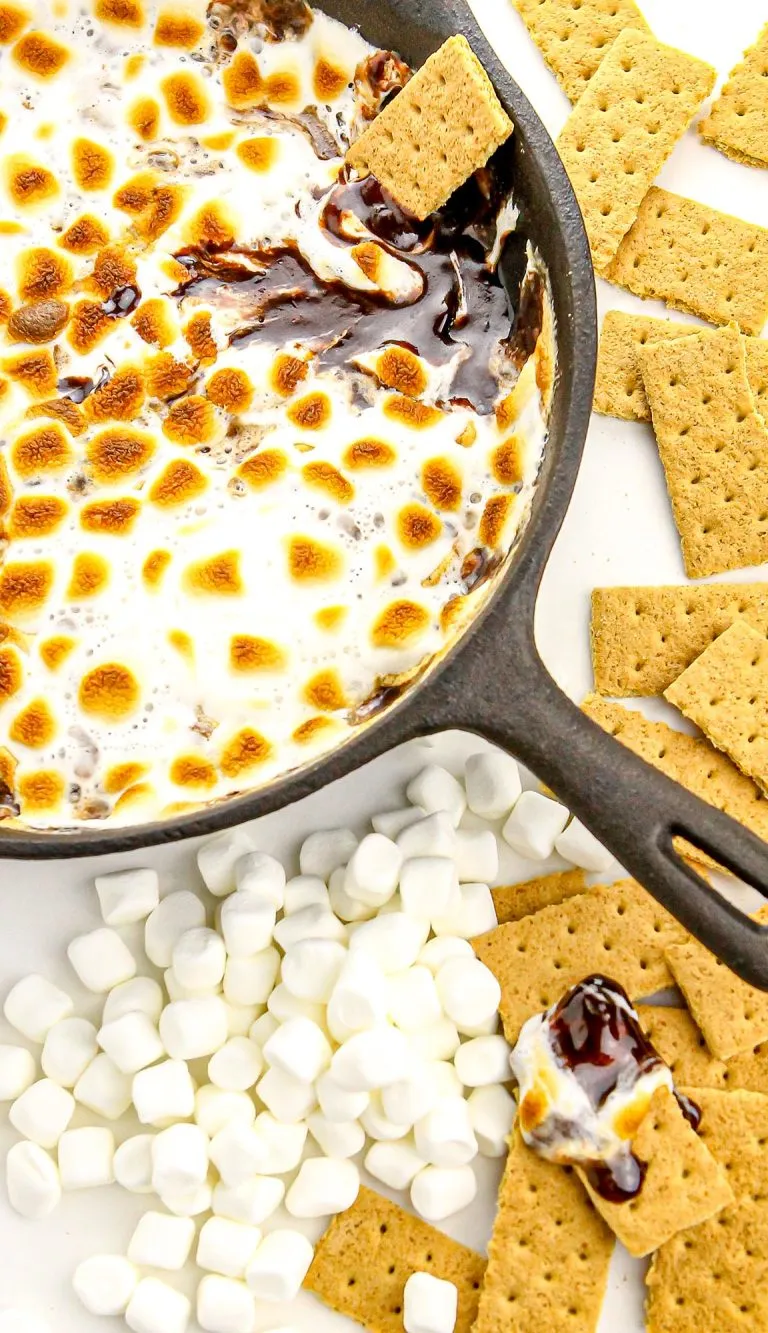 Cast iron skillet with toasted marshmallows and melted chocolate, surrounded by mini marshmallows and graham crackers on a white surface.