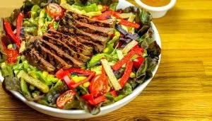 Grilled sliced steak served on a bed of mixed greens, cherry tomatoes, corn, and colorful tortilla strips with dressing on the side.