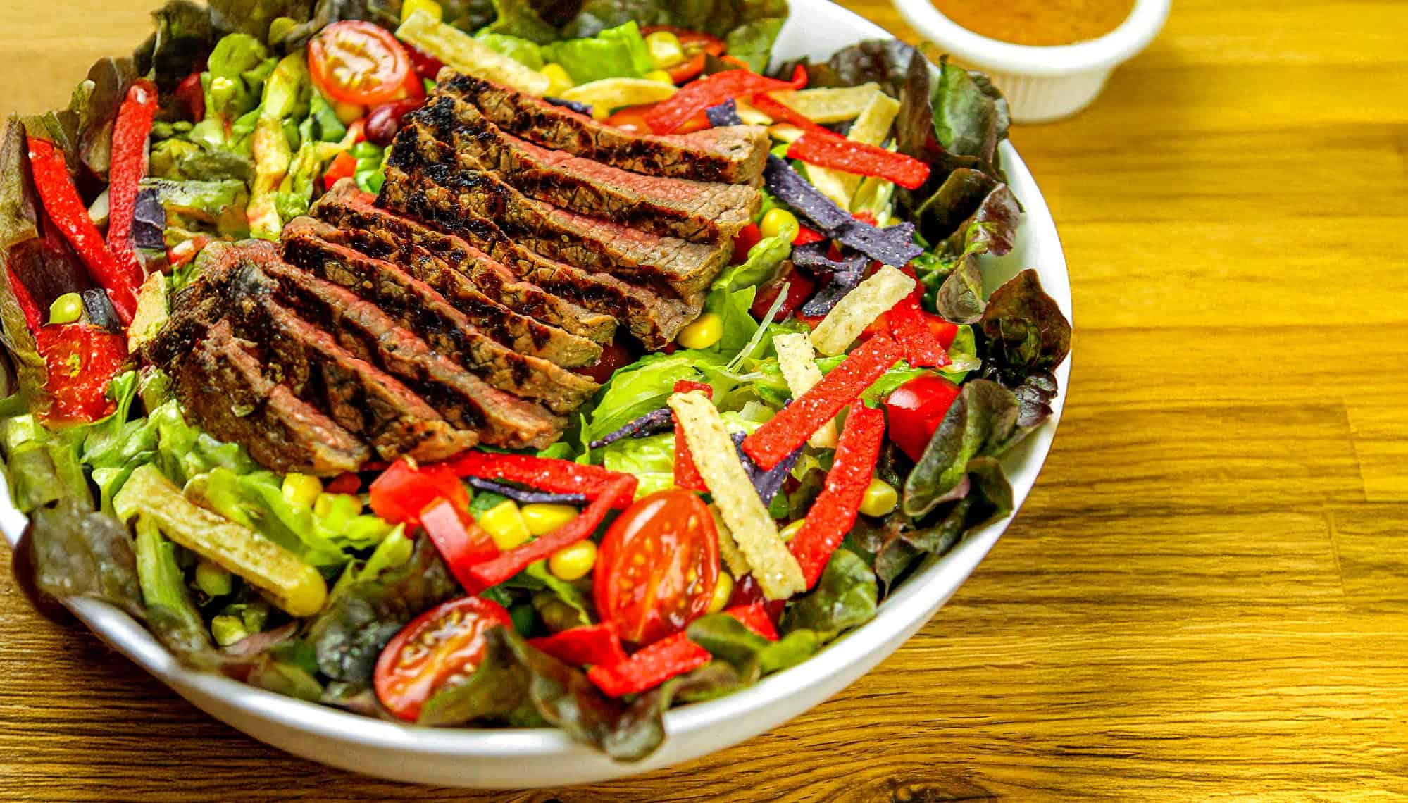 Grilled sliced steak served on a bed of mixed greens, cherry tomatoes, corn, and colorful tortilla strips with dressing on the side.