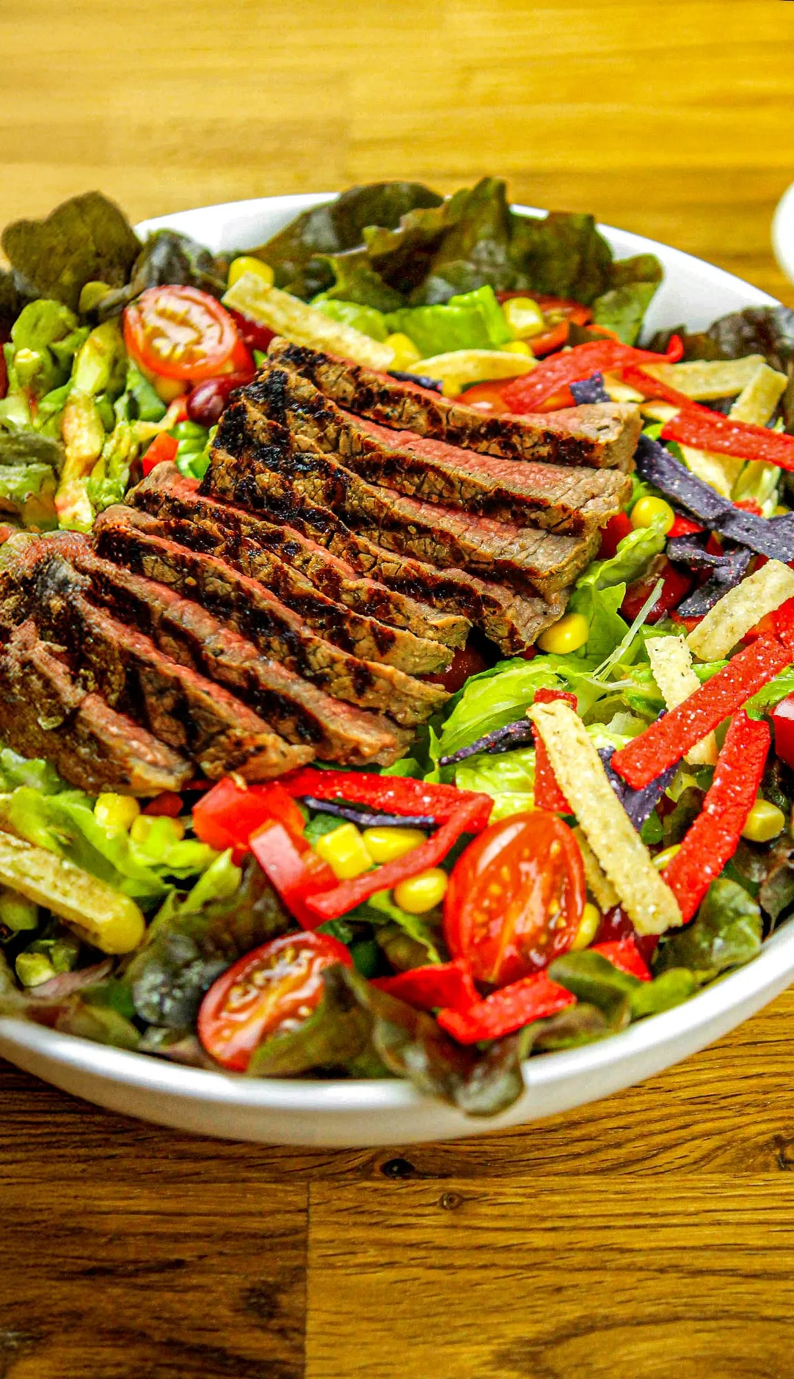 Grilled steak slices on mixed salad with cherry tomatoes, corn, lettuce, and red, purple, and white tortilla strips in a white bowl.