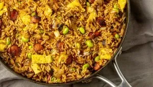Fried rice with scrambled eggs, sausage slices, edamame, and onions in a black skillet on a gray cloth.
