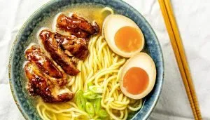 Bowl of ramen with braised pork, soft-boiled eggs, sliced green onions, and broth, next to wooden chopsticks on white cloth.