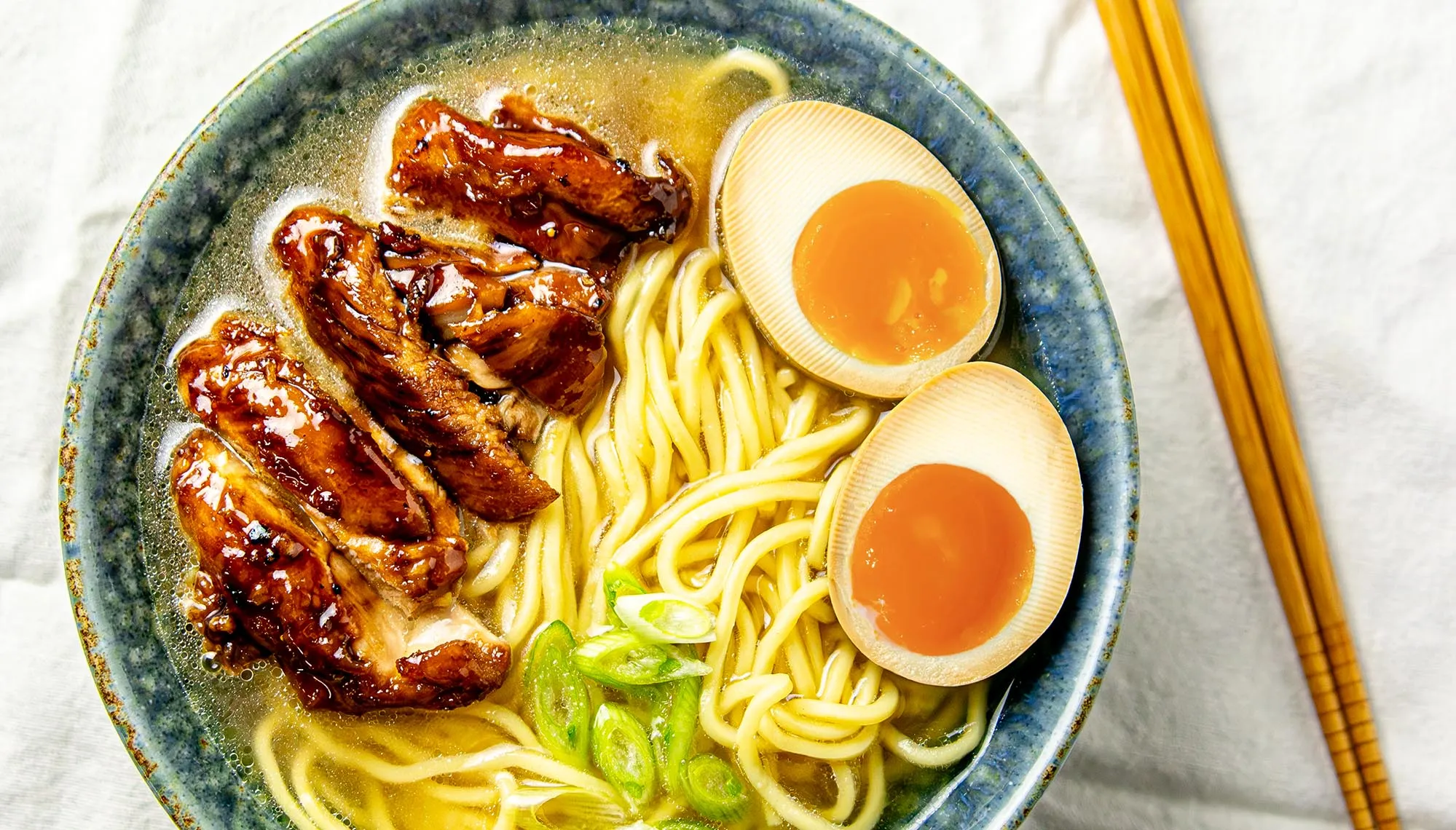 Bowl of ramen with braised pork, soft-boiled eggs, sliced green onions, and broth, next to wooden chopsticks on white cloth.
