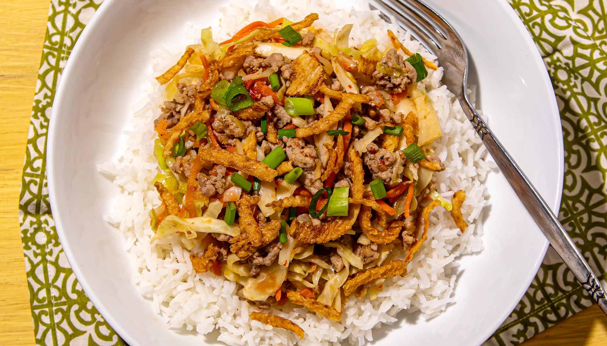 White rice topped with ground meat, shredded cabbage, carrots, fried onions, and chopped green onions in a white bowl with a spoon.