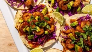 Shrimp tacos with purple cabbage, avocado, cilantro, and lime wedges on small flour tortillas.