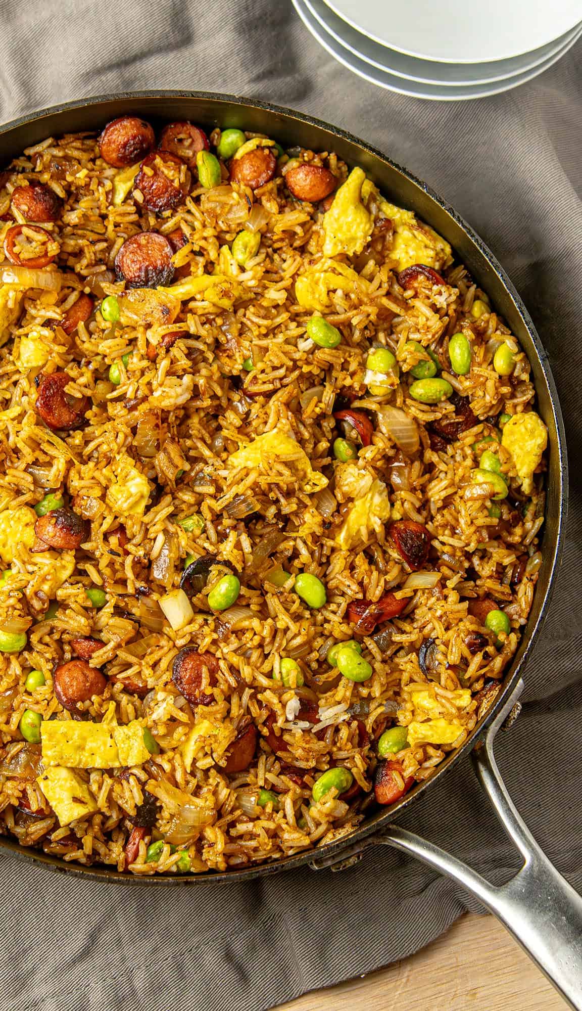 Fried rice with scrambled eggs, sliced sausage, edamame, and onions in a skillet on a gray cloth next to stacked white plates.