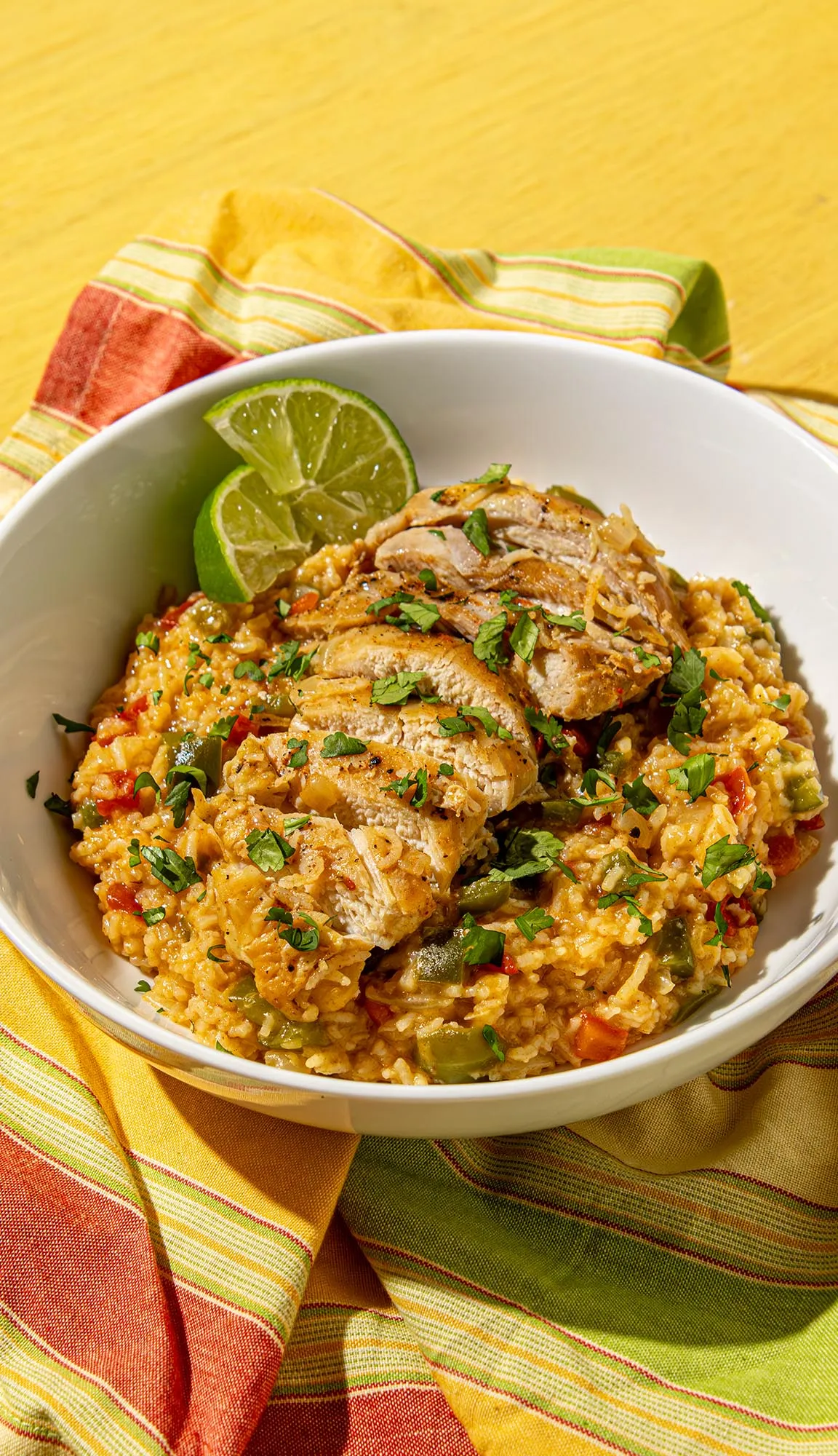 Grilled chicken slices served over seasoned rice with vegetables, garnished with chopped herbs and lime wedges in a white bowl on a colorful napkin.