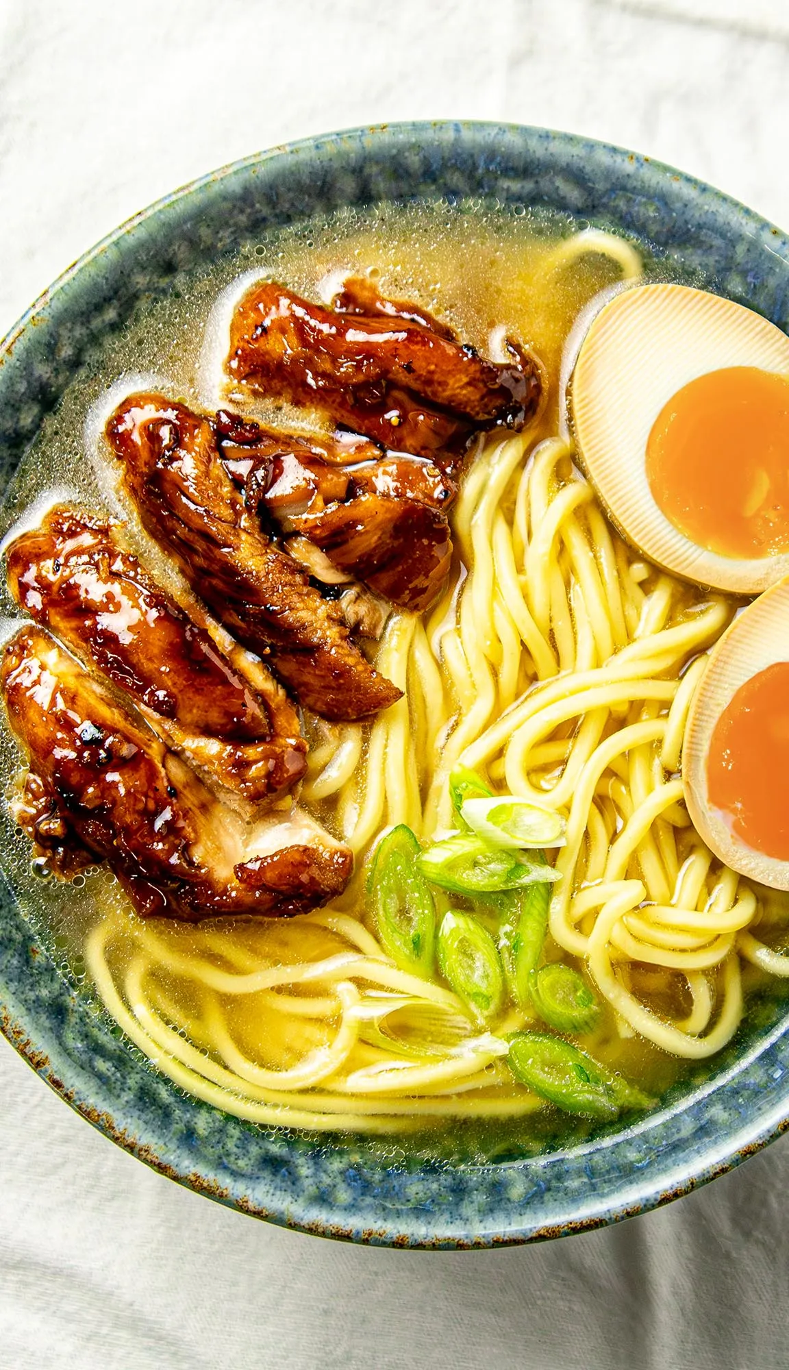 Bowl of ramen with sliced grilled chicken, soft-boiled eggs, green onions, and yellow noodles in clear broth.