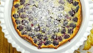 Blackberry clafoutis dusted with powdered sugar in a white scalloped ceramic dish on a wooden table.
