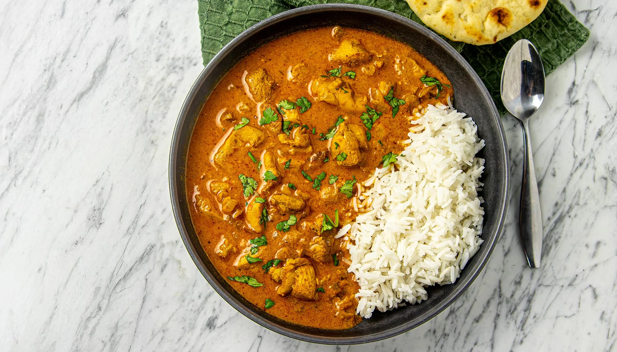 Chicken curry garnished with cilantro served with white rice and naan on a dark plate with a spoon on a marble surface.