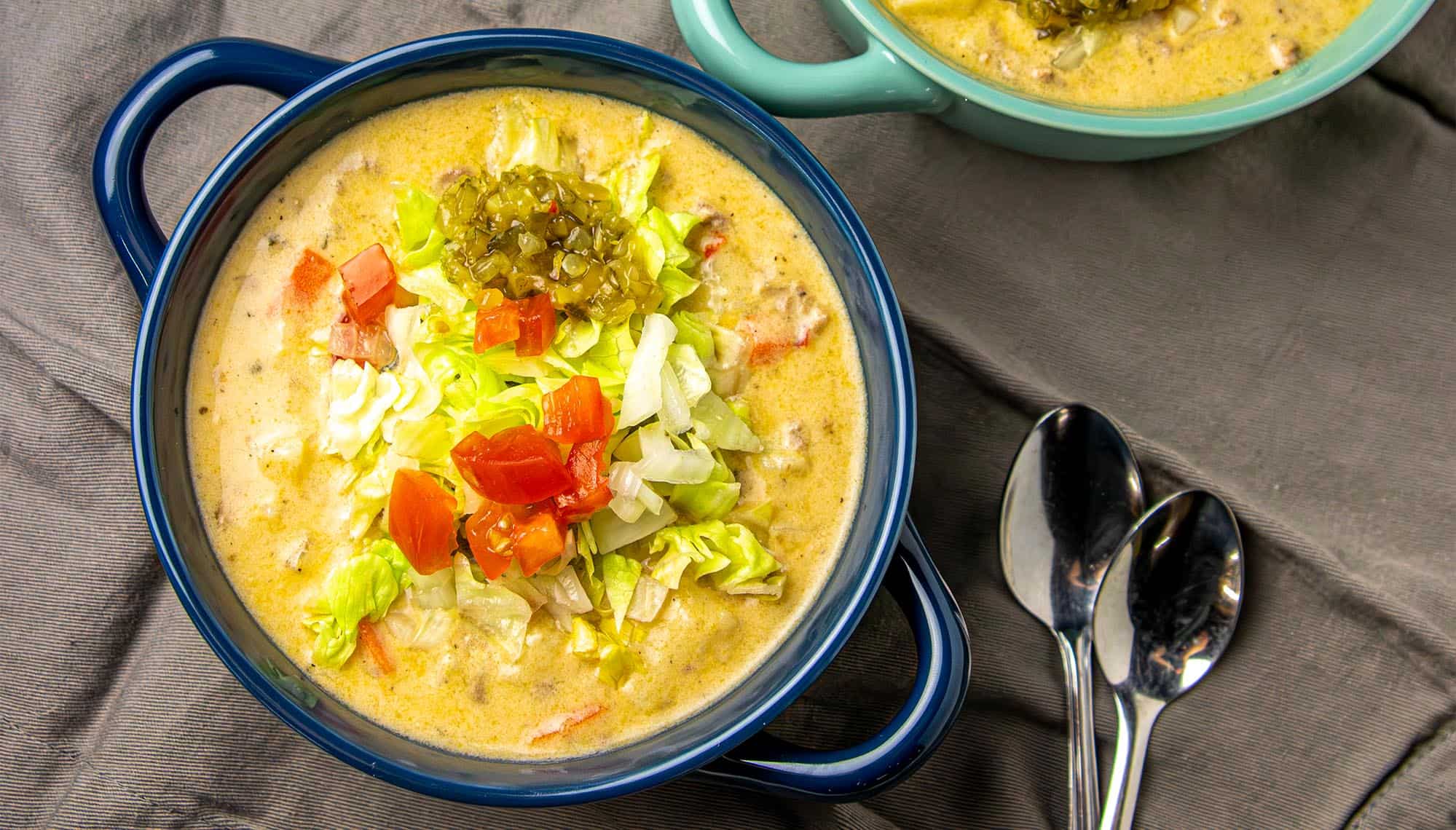 Creamy soup topped with lettuce, diced tomatoes, onions, and relish in a blue bowl with two spoons on a gray cloth.