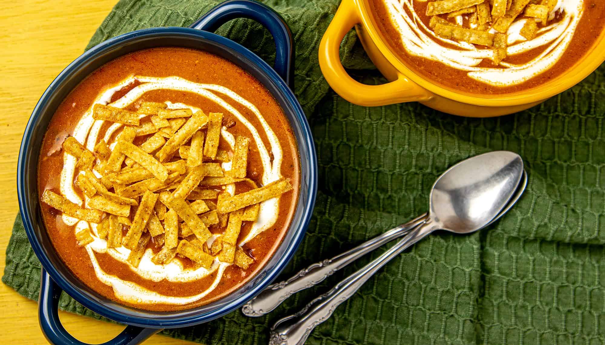 Two bowls of creamy tortilla soup topped with sour cream and crispy tortilla strips, on a green cloth with two silver spoons.