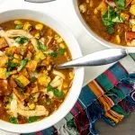 Two bowls of chicken tortilla soup topped with tortilla chips, cheese, corn, tomatoes, and cilantro on a colorful woven placemat.