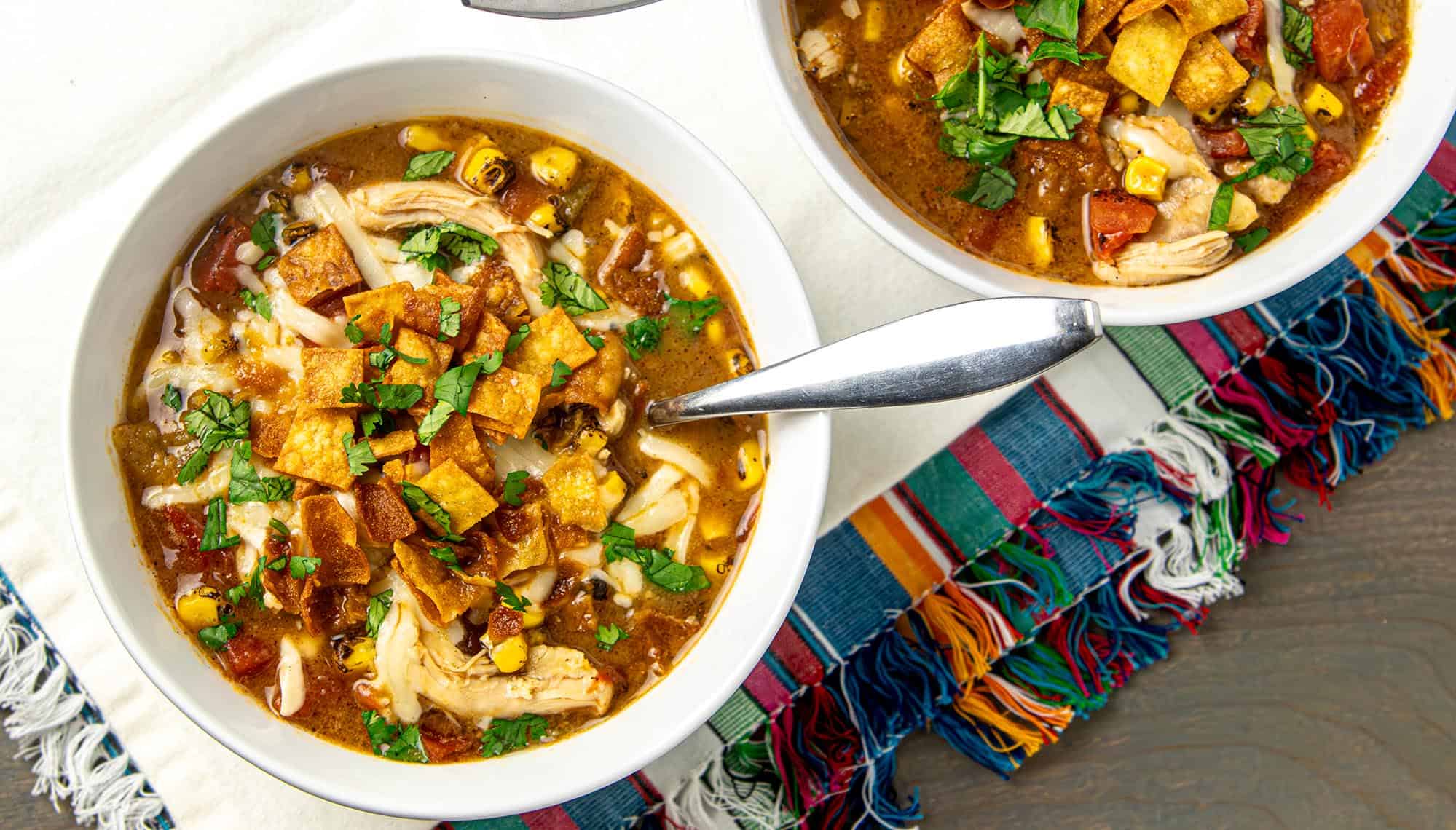 Two bowls of chicken tortilla soup topped with tortilla chips, cheese, corn, tomatoes, and cilantro on a colorful woven placemat.