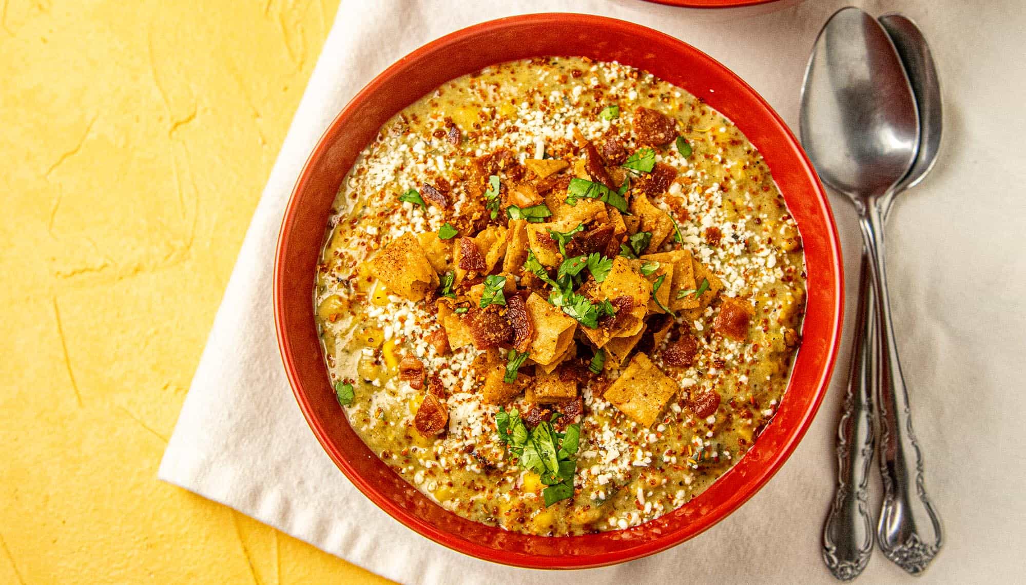 Red bowl with creamy quinoa corn chowder topped with crushed tortilla chips, chopped herbs, and crumbled cheese, on a white cloth with two spoons.