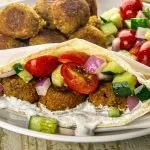 Pita stuffed with falafel, cucumber, cherry tomatoes, red onion, and white sauce, served with extra falafel and salad on a white plate.
