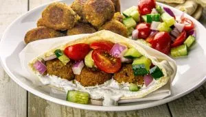 Pita stuffed with falafel, cucumber, cherry tomatoes, red onion, and white sauce, served with extra falafel and salad on a white plate.