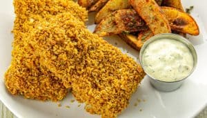 Crispy breaded fish fillets with seasoned potato wedges and a cup of tartar sauce on a white plate.
