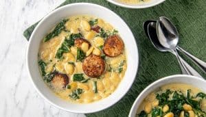 Bowl of creamy white bean soup with kale and browned sausage slices, next to two spoons on a green textured napkin.