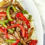 Beef stir-fry with red and green bell peppers and onions over white rice on a white oval plate with a striped cloth background.