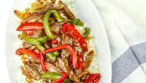 Beef stir-fry with red and green bell peppers and onions over white rice on a white oval plate with a striped cloth background.