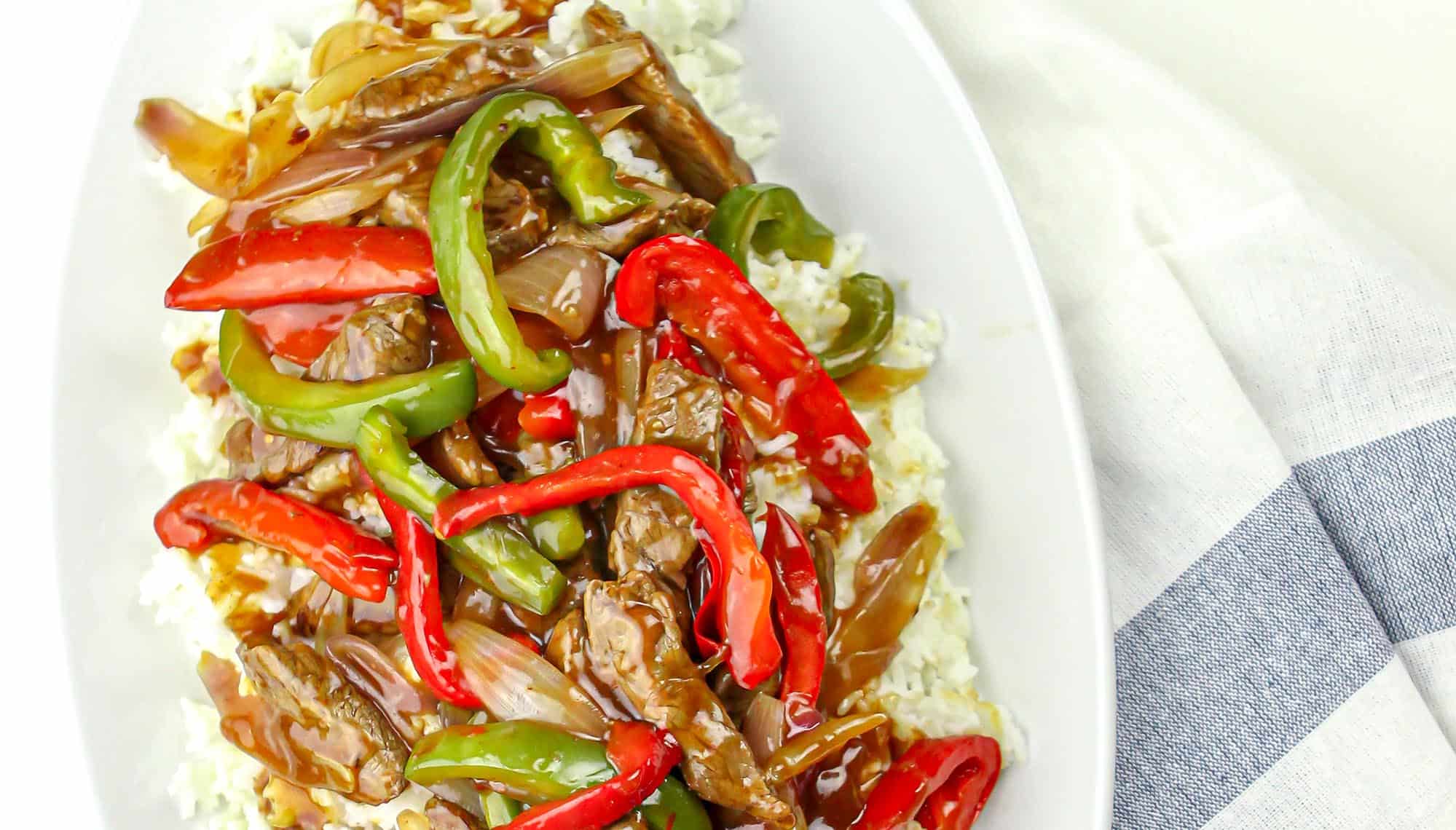 Beef stir-fry with red and green bell peppers and onions over white rice on a white oval plate with a striped cloth background.