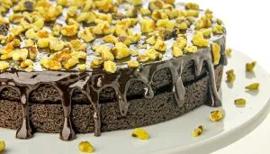 Chocolate cake with glossy chocolate ganache dripping down sides, topped with chopped walnuts on a white plate.