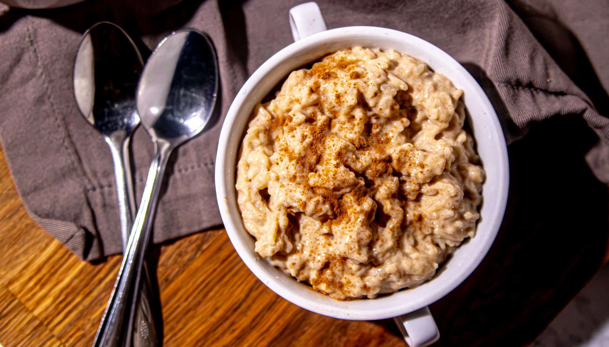 Creamy rice pudding sprinkled with cinnamon in white mug next to two spoons on brown cloth on wooden surface.