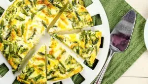 Asparagus and cheese frittata cut into slices on a white serving platter with a silver spatula on a green cloth.