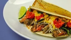Steak taco with grilled cherry tomatoes, corn, shredded cheese, and greens on a white plate with a lime wedge.