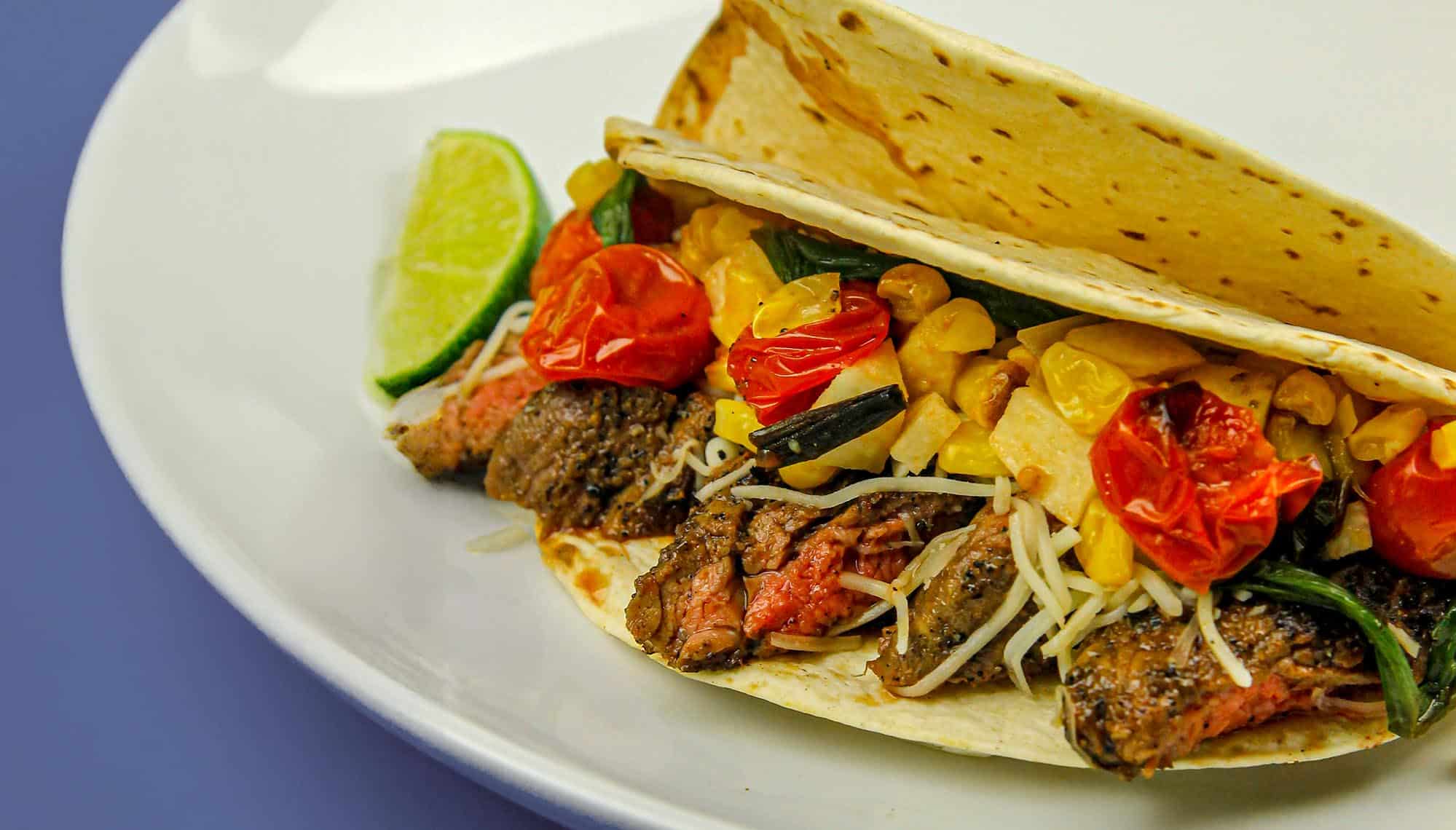 Steak taco with grilled cherry tomatoes, corn, shredded cheese, and greens on a white plate with a lime wedge.