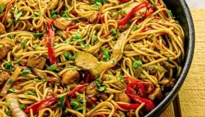 Stir-fried noodles with chicken, red bell peppers, onions, and chopped green herbs in a black skillet on a yellow cloth.