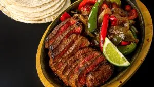 Grilled sliced steak with sautéed bell peppers and onions, garnished with a lime wedge, served with warm tortillas on the side.